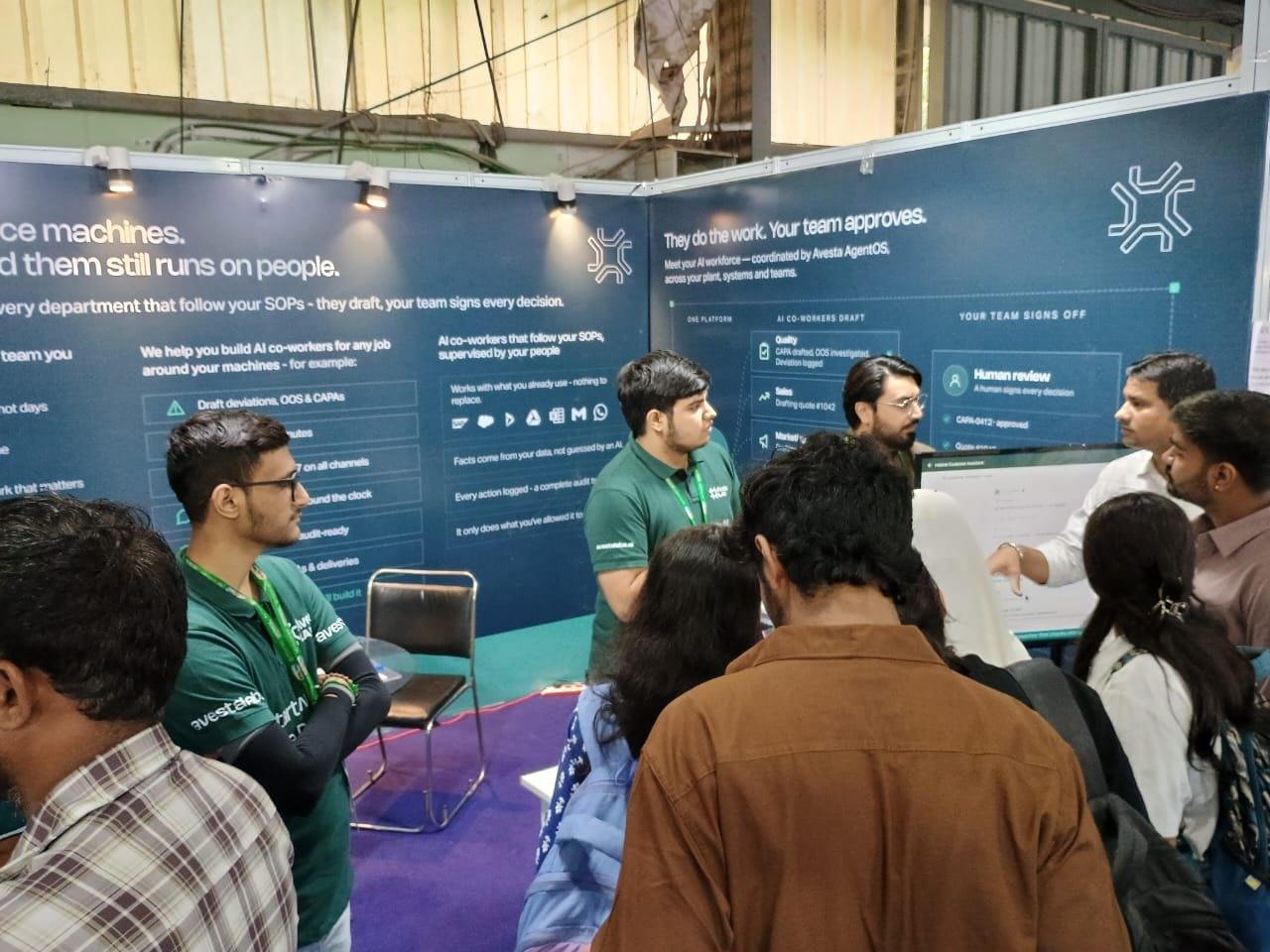 An Avesta Labs engineer pointing out a human review step on the demo screen to a group of visitors, under a wall panel reading they do the work, your team approves